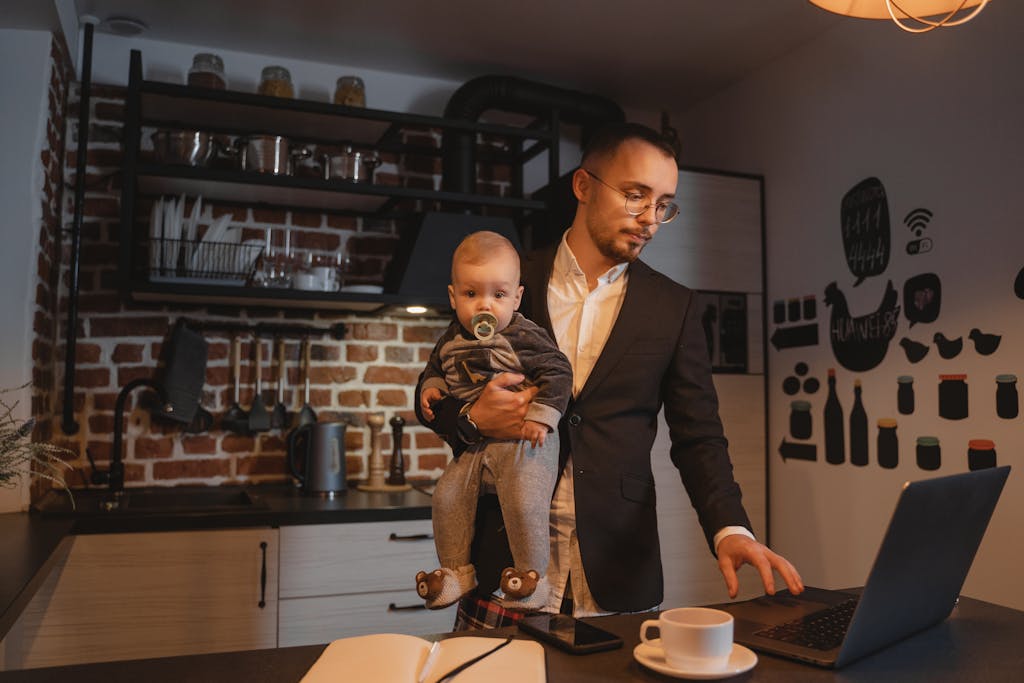 Father working on a laptop while holding his baby in a kitchen setting.